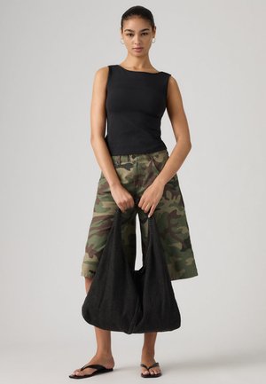 Donna con top nero senza maniche, pantaloncini mimetici fino al ginocchio, infradito nere, che tiene in entrambe le mani una grande borsa nera morbida.