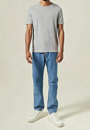 Homme portant un t-shirt à manches courtes gris clair uni, un jean bleu et des baskets blanches, debout devant un fond beige.