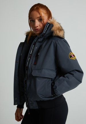 Chaqueta aislante gris con capucha forrada de piel, cremallera con botones a presión, bolsillos laterales y parche de logo en la manga.