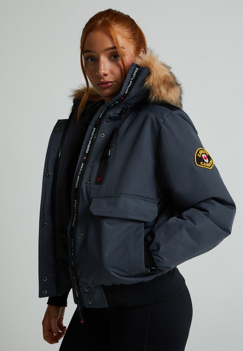 Pelēka izolēta jaka ar kažokādas apšuvumu uz kapuces, rāvējslēdzējs ar pogām, sānu kabatas un logo plāksteris uz piedurknes.