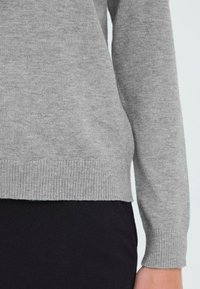 Grå strikkegenser med ribbet mansjetter og kant. Myk tekstur og tettsittende design, kombinert med et svart plagg under.