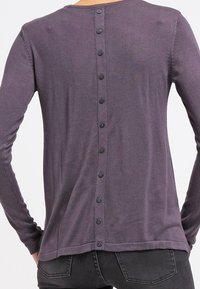 Lila långärmad tröja med rund halsringning och en rad svarta knappar på ryggen. Mjuk stickad tyg med en slät textur.