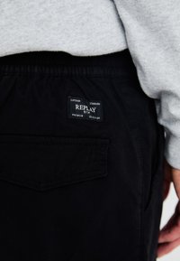 Svart bukser med en baklomme som har et rektangulært merke som sier "REPLAY." Materialet ser mykt ut med en matte tekstur.