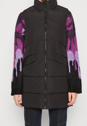 Veste matelassée noire sans manches avec fermeture éclair à l'avant, portée par-dessus un pull à manches longues présentant des motifs abstraits en noir, violet et rose.