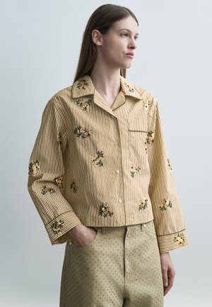 Naine, kellel on pikad pruunid juuksed, kannab beeži ja valge triibulist särki lillelise tikandiga ning helerohelisi täpilise mustriga pükse.