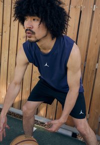 Tengeri kék ujjatlan felső és fekete rövidnadrág fehér Jumpman logóval, izmos testalkat, kosárlabdázik, fa paneles háttér.