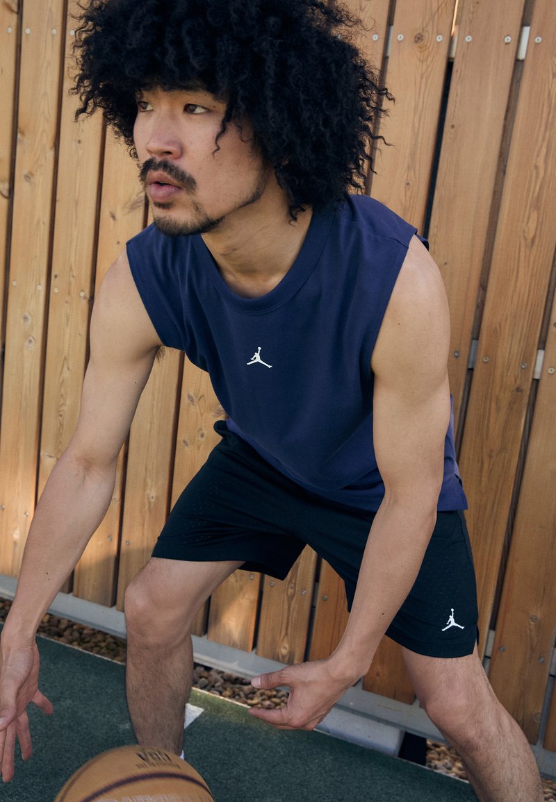 Tengeri kék ujjatlan felső és fekete rövidnadrág fehér Jumpman logóval, izmos testalkat, kosárlabdázik, fa paneles háttér.
