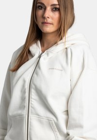 Bílá mikina na zip s měkkou texturou, vybavená prostornou kapucí a předními kapsami. Logo "PEGADOR" je diskrétně umístěno na hrudi.