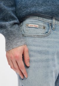 Γκρίζα ζακέτα από μείγμα μαλλιού με ραμμένο μανίκι, συνδυασμένη με ανοιχτό μπλε τζιν παντελόνι που διαθέτει μια κόκκινη ετικέτα "Calvin Klein" κοντά στην πίσω τσέπη.