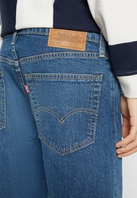 Jean en denim bleu avec une pièce en cuir à la taille, des coutures courbes sur les poches arrière, et une étiquette rouge Levi's sur la poche gauche.