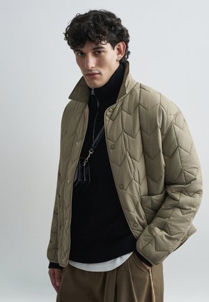 Veste beige matelassée avec un col montant, dotée d'une fermeture à boutons et d'une couche intérieure noire avec zip. Portée sur un pantalon sombre.
