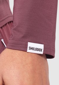 Maniche atletiche bordeaux, realizzate in tessuto texturizzato, presentano un'etichetta bianca con la scritta "SMILODOX" al polsino. Vista ravvicinata dell'orlo.