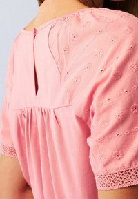 Blouse rose avec des motifs floraux brodés sur les manches, une fermeture à clé dans le dos et des détails de tissu froncé à la taille.