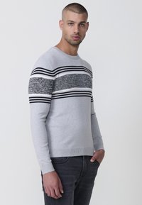 Suéter gris con rayas horizontales en blanco y negro y un diseño texturizado en el pecho. Mangas largas y cuello redondo.
