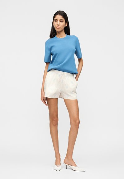 Femme debout portant un haut bleu à manches courtes, un short blanc à motifs floraux et des mules blanches à talons, sur un fond clair uni.