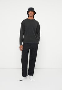 Crna košulja dugih rukava i široke crne hlače, uz crnu šeširicu. Bijele tenisice upotpunjuju odjevnu kombinaciju, postavljenu prema jednostavnoj pozadini.