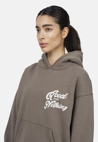 Bruine hoodie met een voorzak in kangoeroestijl, met witte tekst "Good for Nothing" in een gestileerd lettertype. Gemaakt van zachte, gestructureerde stof.