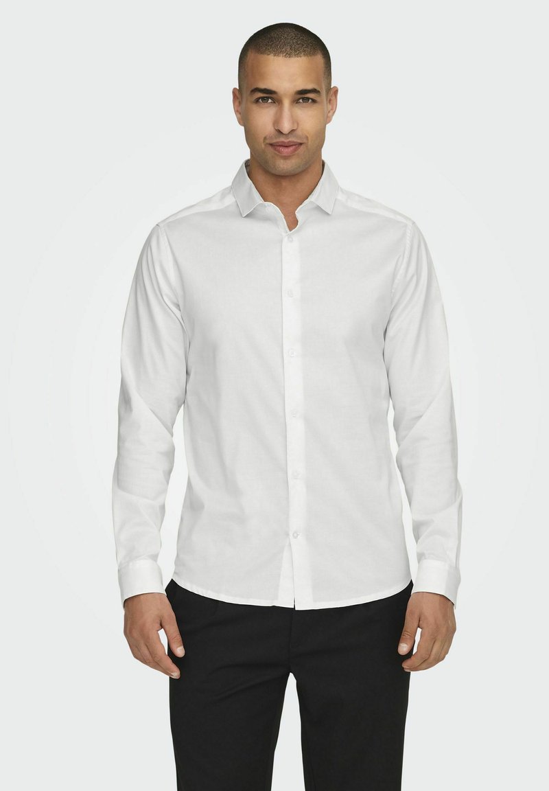 Chemise blanche à manches longues en tissu lisse, avec un col classique et une coupe ajustée, associée à un pantalon sombre.