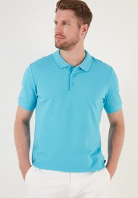 Camisa polo azul claro hecha de tejido texturizado, con cuello clásico, mangas cortas y una tapeta de tres botones. Se lleva pantalones blancos.