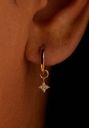 Goldener Creolen-Ohrring mit einem kleinen, sternförmigen Anhänger, besetzt mit klaren Steinen, getragen an einem dunkelhäutigen Ohr.