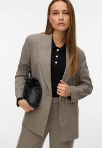 Blazer grigio con rever affilati, indossato sopra una polo nera a costine con tre bottoni decorativi bianchi. Clutch nera a trama appuntita tenuta in mano.