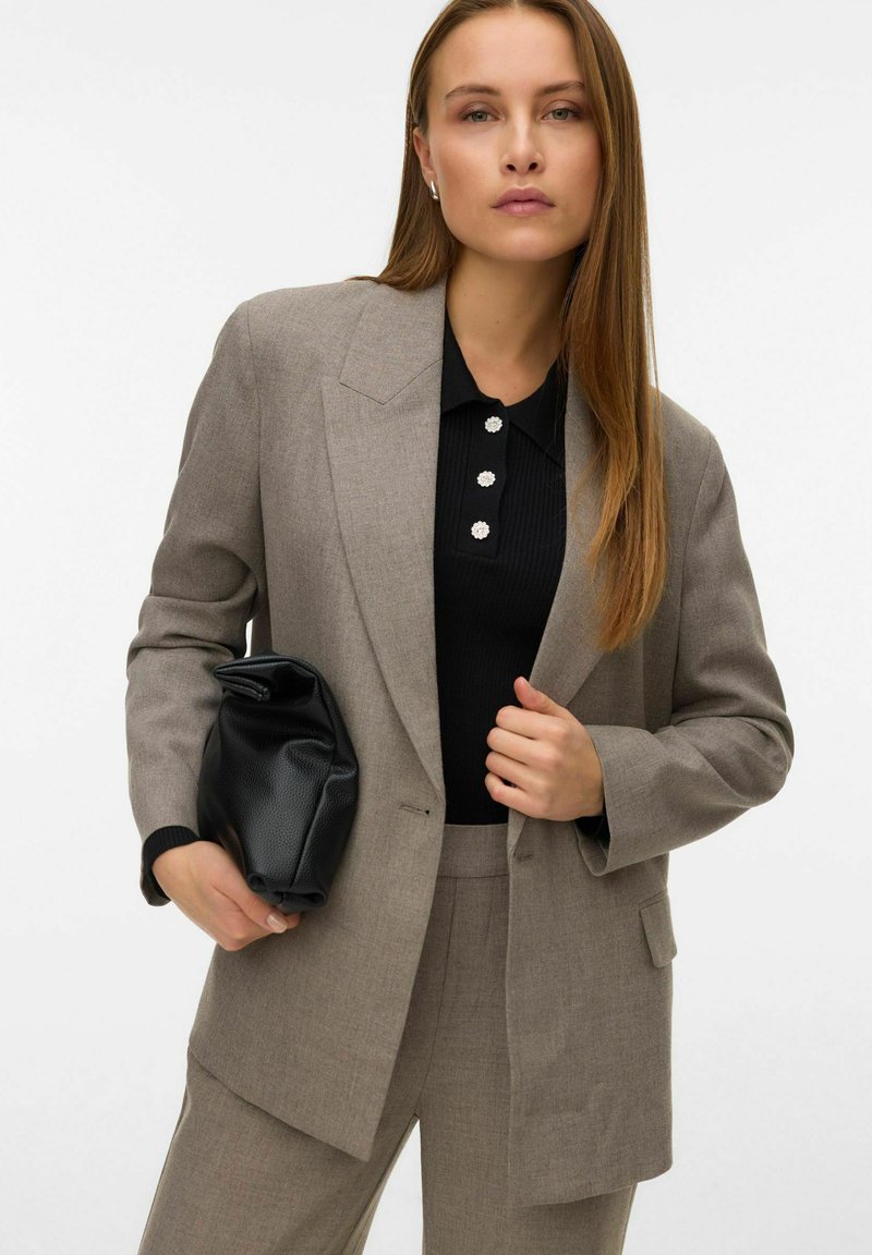 Blazer grigio con rever affilati, indossato sopra una polo nera a costine con tre bottoni decorativi bianchi. Clutch nera a trama appuntita tenuta in mano.