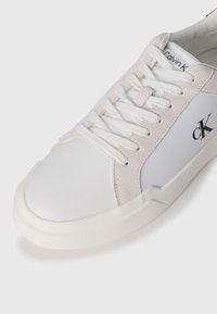 Calvin Klein sneaker met een bovenwerk van wit leer en lichtgrijze suède accenten, ronde neus, platte witte veters en een getextureerde rubberen zool.