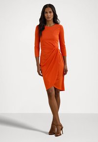 Vestido envolvente laranja com mangas de três quartos, detalhe franzido na cintura e comprimento até ao joelho. Tecido liso e elástico, combinado com sapatos de salto castanhos.