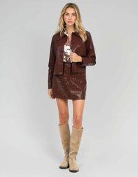 Femme portant une veste en cuir marron et une mini jupe cloutée assortie, un chemisier à motifs et des bottes en suède beiges jusqu'aux genoux se tenant sur un fond gris.