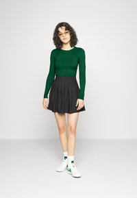 Grön, ribbad långärmad topp som är kombinerad med en svart veckad kjol och vita sneakers med gröna detaljer. Modellen bär gulaktiga glasögon.
