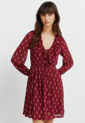 Kvinde iført en langærmet, knælængde, mørkerød kjole med små mønstredeprint og en flæset V-hals med bindedetalje.