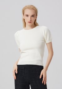 Pull en tricot blanc à manches courtes avec un col rond, présentant des manches bouffantes et un ourlet côtelé ajusté. Texture lisse, sans motifs.