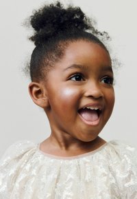 Enfant à la peau foncée et aux cheveux bouclés, portant une robe légère et transparente ornée de paillettes argentées. Elle affiche une expression joyeuse et des yeux pétillants.