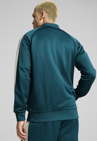 Teal idrottsjacka med en mjuk textur, med krage och kontrasterande beige sidopaneler. Fitted mudd och nederkant. Reflekterande detaljer.