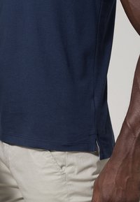 Gros plan d'une personne portant une chemise bleu marine et un pantalon beige clair, montrant la fente latérale de la chemise et le bras droit de la personne.