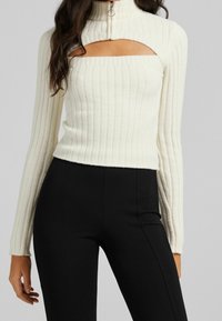 Vit ribbad stickad tröja med hög krage, med en fyrkantig utskärning vid bröstet, i kombination med svarta åtsittande byxor. Dragkedjedetalj vid halsen.