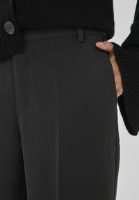 Persona valkā melnas bikses un melnu džemperi, ar vienu roku ieliktu labās bikšu kabatas iekšpusē.