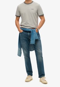 Grijze T-shirt met korte mouwen en donkere afwerking, blauwe spijkerbroek en een lichtblauw overhemd om de taille geknoopt. Off-white sneakers maken de look compleet.