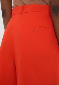 Pantalon rouge sur mesure avec une texture lisse, une ceinture fine, des poches latérales et un bouton décoratif sur la poche arrière.