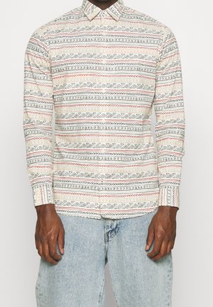 Homme portant une chemise à manches longues boutonnée avec des motifs géométriques horizontaux dans des couleurs sourdes et un jean bleu clair.