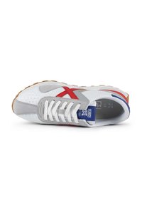 Zapatilla deportiva con una parte superior perforada blanca, acentos en ante gris, detalles rojos y una suela de goma marrón. Presenta cordones blancos y una etiqueta azul.