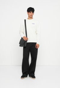 Chemise blanche à manches longues avec logo bleu, associée à un pantalon noir ample et un sac messager noir. Des chaussures décontractées complètent le look.