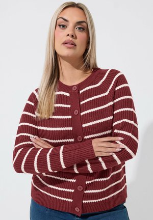Kvinde med langt blond hår iført en vinrød og hvid stribet knaptrøje, stående med armene krydset mod en ensartet baggrund.