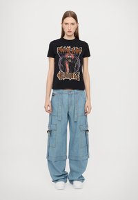 Μαύρο βαμβακερό T-shirt με γραφικά τίγρης και κείμενο, συνδυασμένο με ανοιχτό γαλάζιο παντελόνι cargo σε φαρδιά γραμμή με τσέπες που κουμπώνουν και ασημένια εξαρτήματα.