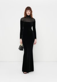 Black velvet long-sleeve dress with a high mesh neckline, fitted silhouette, and floor-length design. Accented with a small black handbag.
