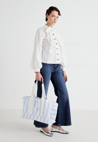 Mulher vestindo uma blusa branca com babados e jeans azul boca de sino, segurando uma grande bolsa de mão listrada em azul e branco, de pé sobre um fundo branco.