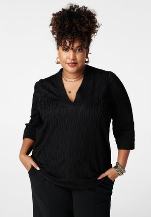 Vrouw met krullend haar, gekleed in een zwarte top met textuur en zwarte broek, geaccentueerd met gouden creolen, meerdere kettingen en armbanden, met handen in de zakken.