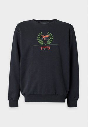 Jeune homme debout, une main dans la poche, portant un sweat-shirt noir avec un logo rouge et vert, un pantalon foncé, des chaussettes blanches et des baskets noires.