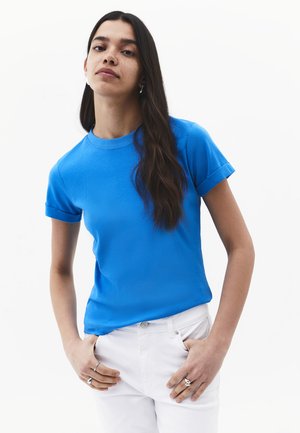 Frau mit langen dunklen Haaren, die ein leuchtend blaues Kurzarmshirt und weiße Hose trägt, steht mit den Händen in den Taschen vor weißem Hintergrund.