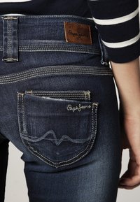 Mörkblå denimjeans med en brun läderetikett. Har två bakfickor, varav en med vit söm och "Pepe Jeans" broderi.
